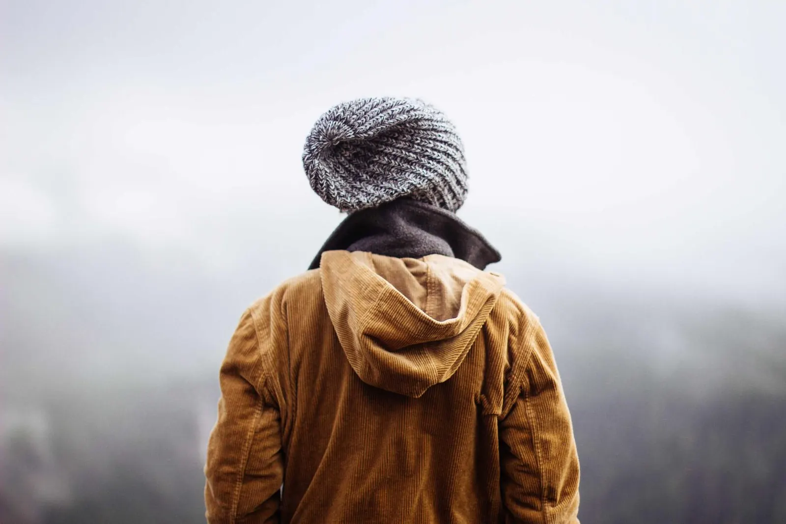 Jovem de costas, com gorro e casaco caramelo, olhando para uma paisagem enevoada
